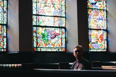 A man sits alone in a church with stained glass windows